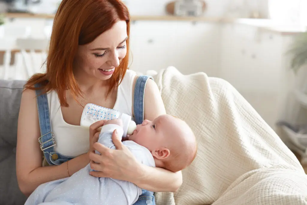 portrait of happy tender young mother with ginger hair feeding her baby boy from milk bottle. beautiful mom feeds infant son with formula while sitting on couch at home. motherhood and happiness